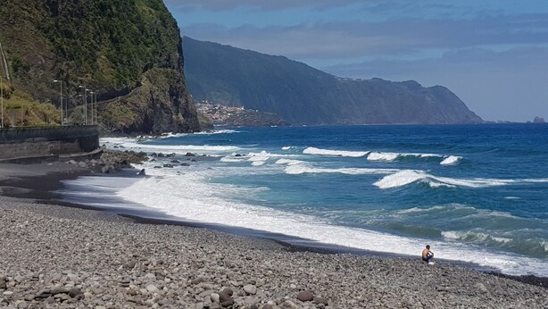 A rocky beach with waves crashing onto the shore, a person sitting on the rocks, and a mountainous backdrop.