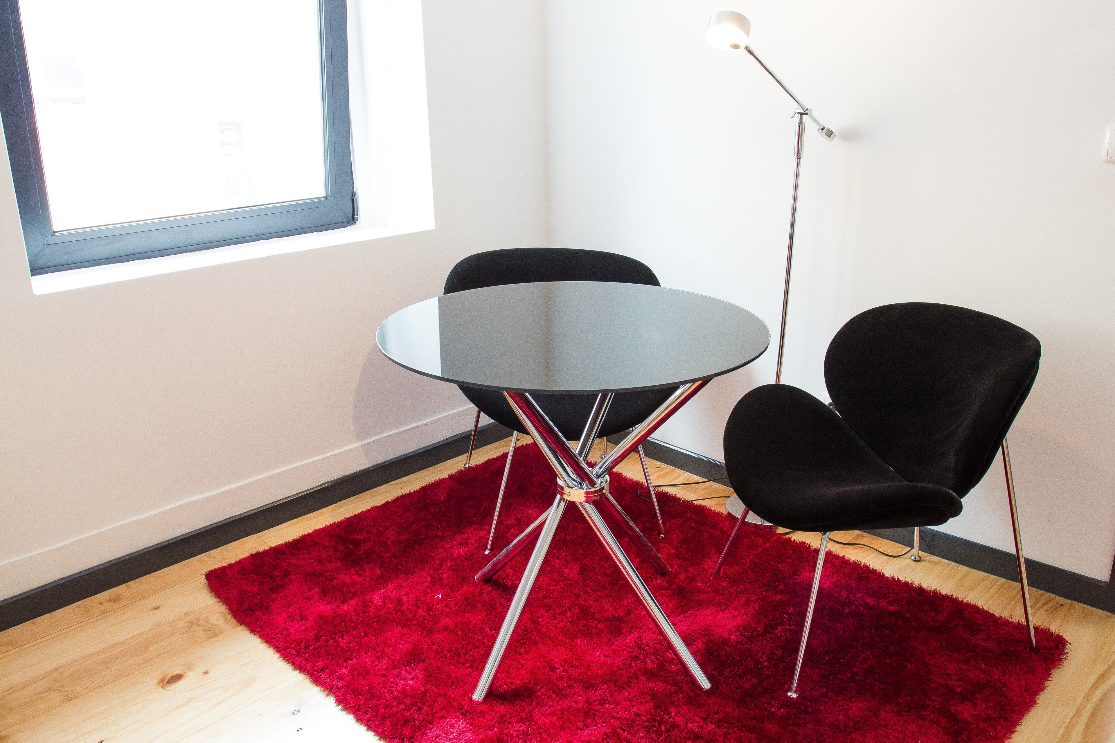 A room with a round table, two black chairs, a red rug, a floor lamp, and a window.