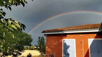 A house with a red exterior and a white door, a small patio with a table and chairs, and a rainbow in the sky.
