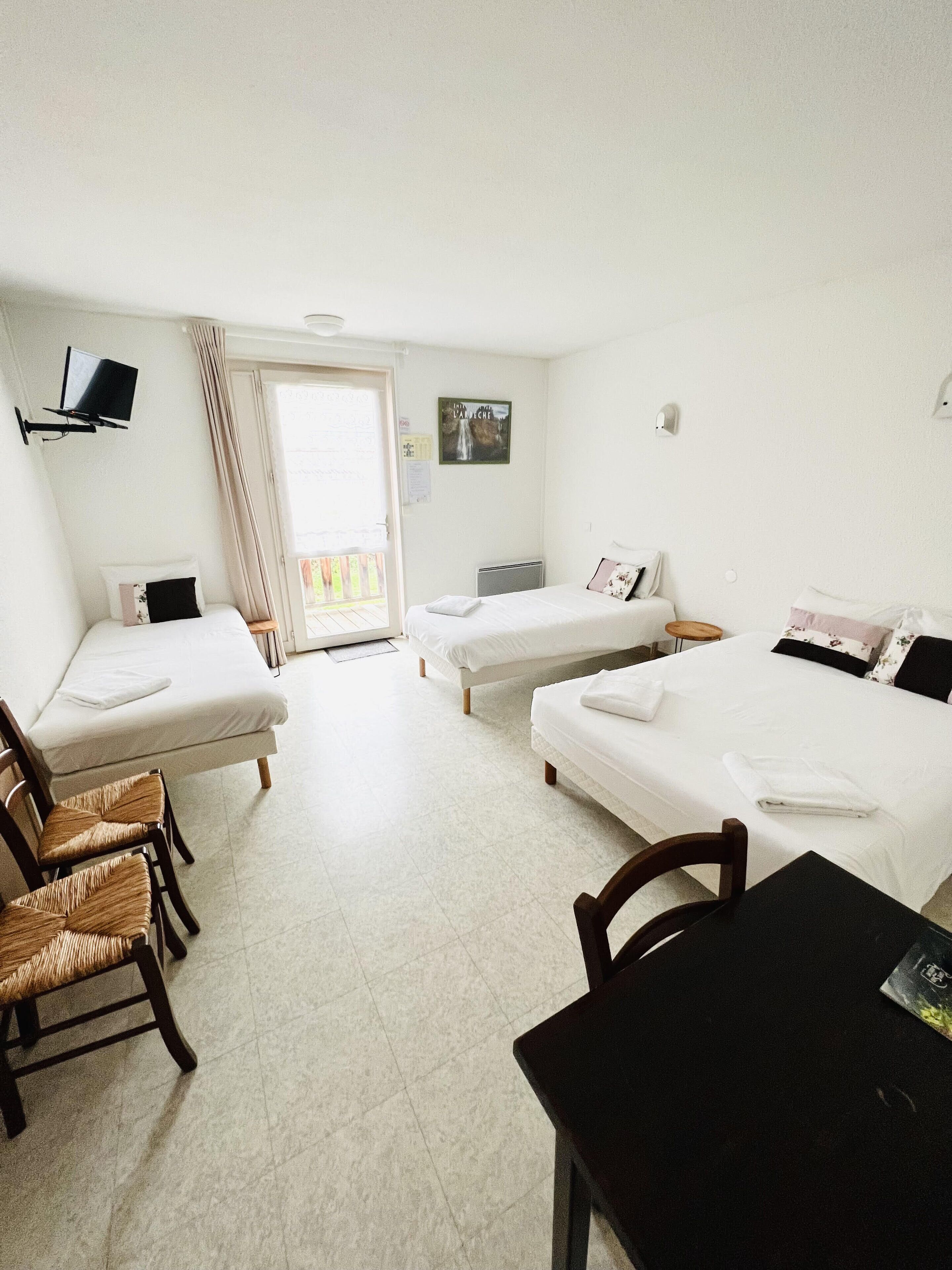 A hotel room with two single beds, a small table, two wicker chairs, a TV mounted on the wall, and a balcony with a view.
