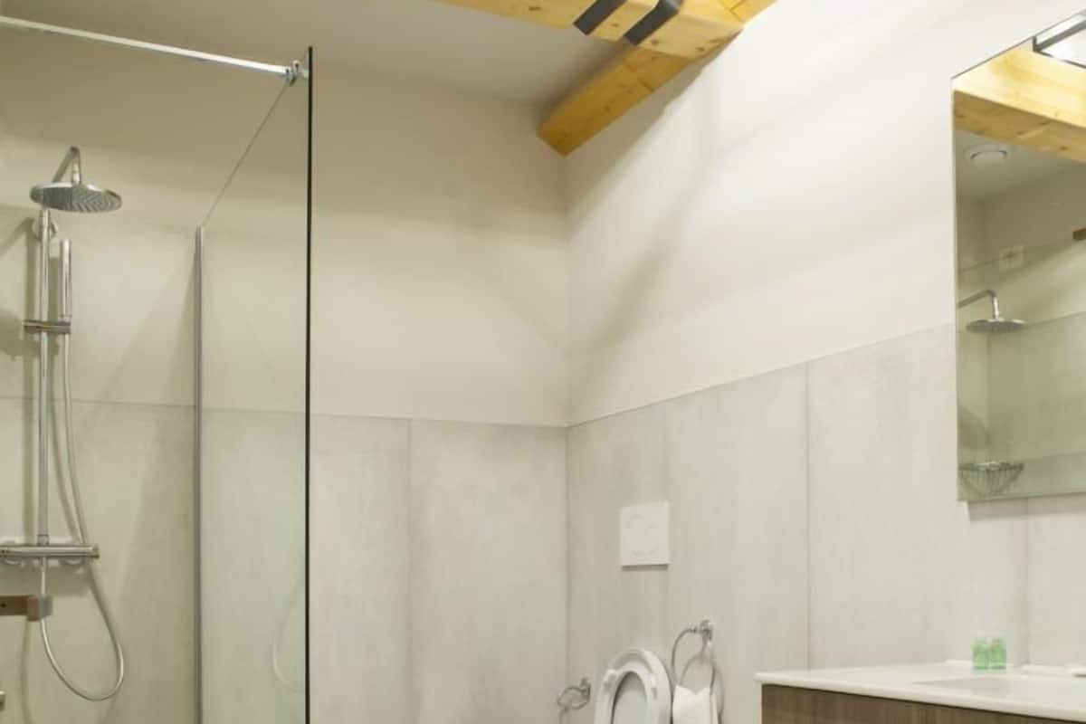 A modern bathroom with a glass-enclosed shower, two toilets, a vanity with a mirror, and a wooden ceiling beam.