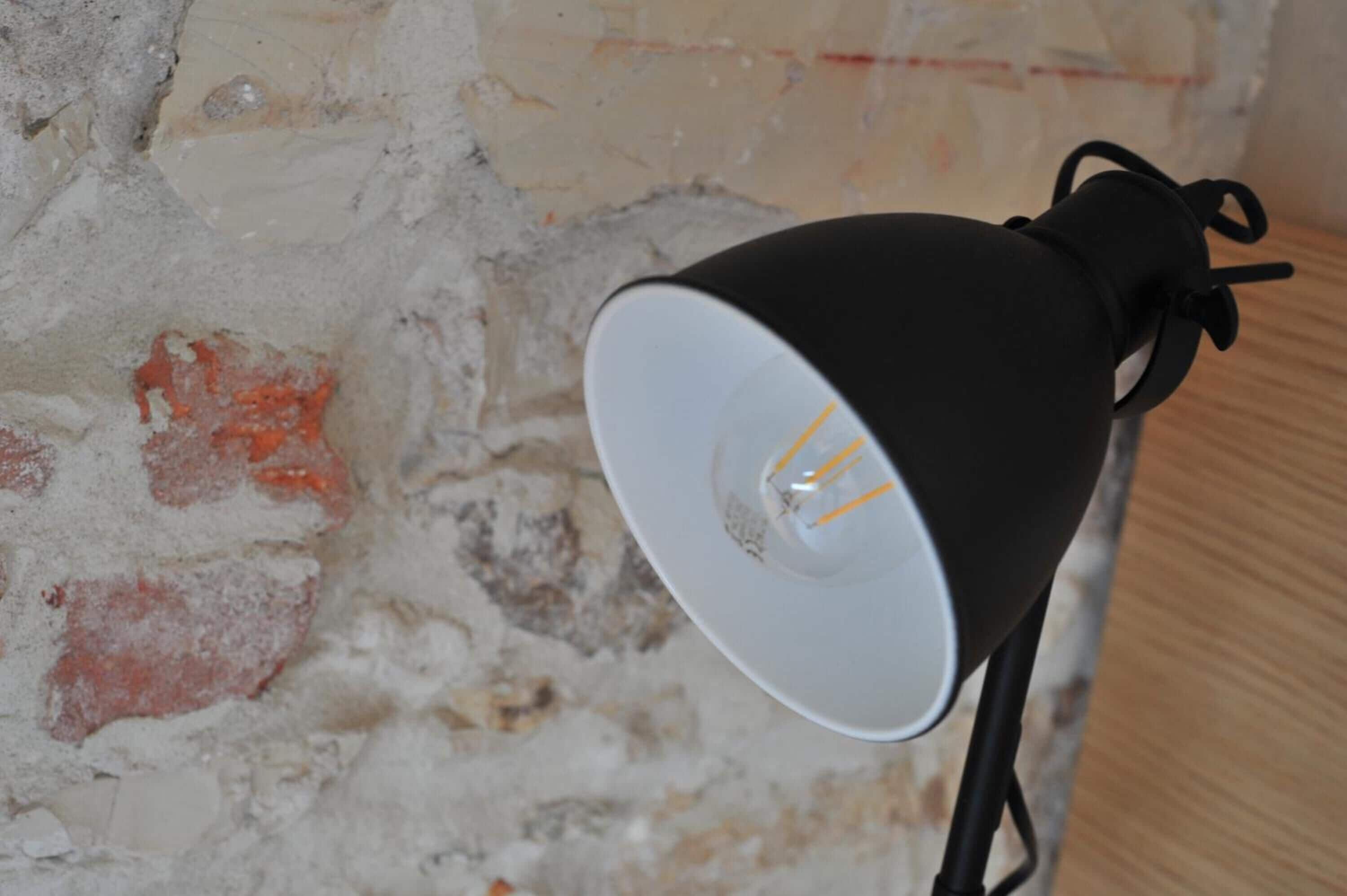 Una lampada nera con paralume bianco davanti a un muro strutturato con mattoni a vista.