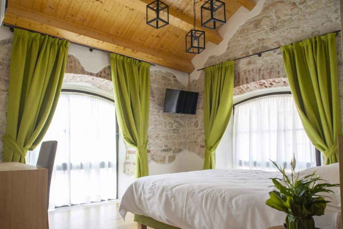 A bedroom with a large bed, green curtains, a flat-screen TV, and a wooden ceiling.
