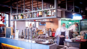 Un espace bar avec un comptoir, des étagères remplies de bouteilles et une cuisine en arrière-plan.