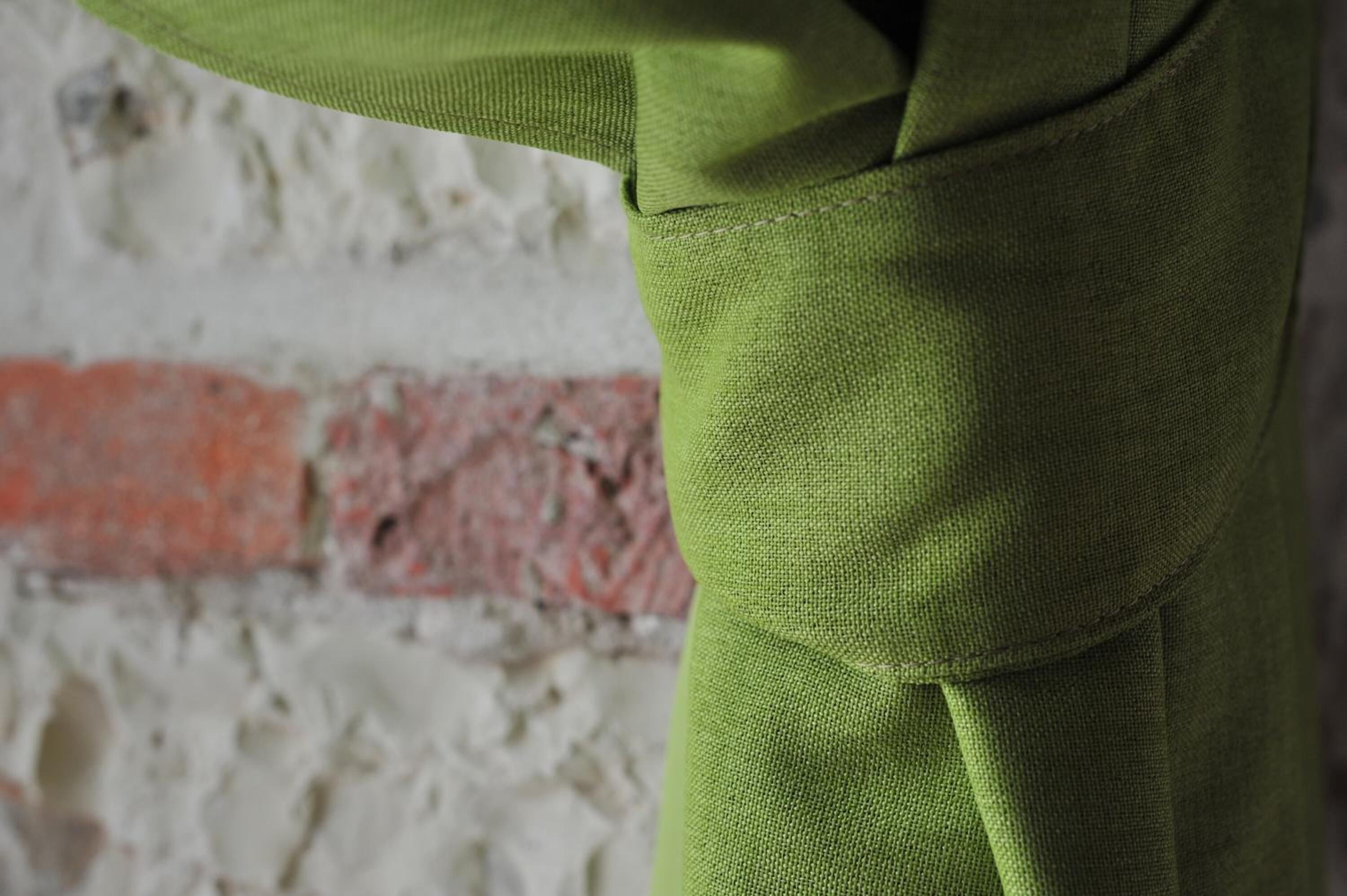 Un gros plan d’un tissu vert à la texture particulière, qui pourrait être un morceau de vêtement ou un revêtement d’ameublement, sur un fond constitué d’un mur brut et non fini, avec un motif de briques rouges.