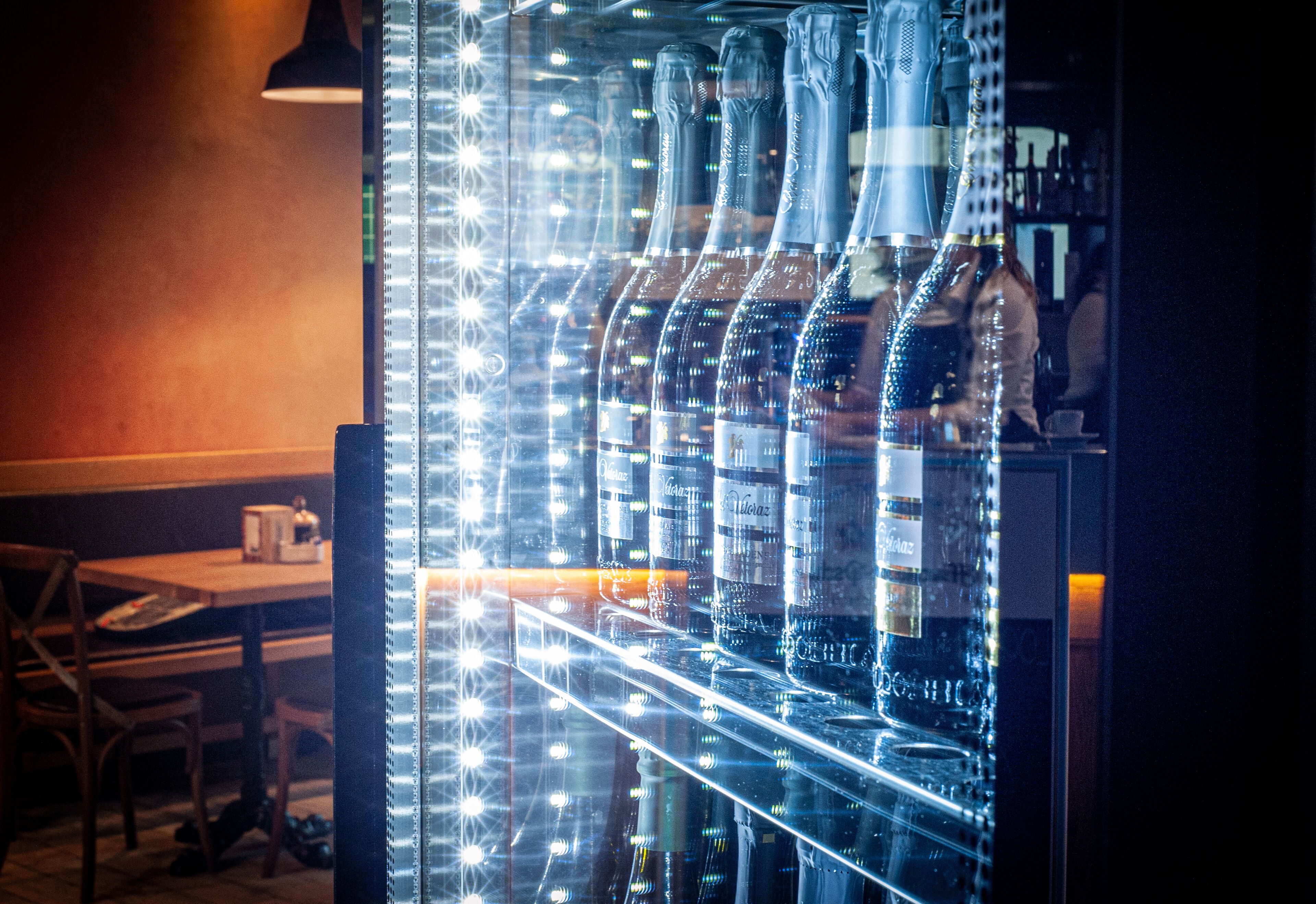 Zona de bar con una vitrina de vidrio que contiene botellas, una mesa con sillas y una persona al fondo.