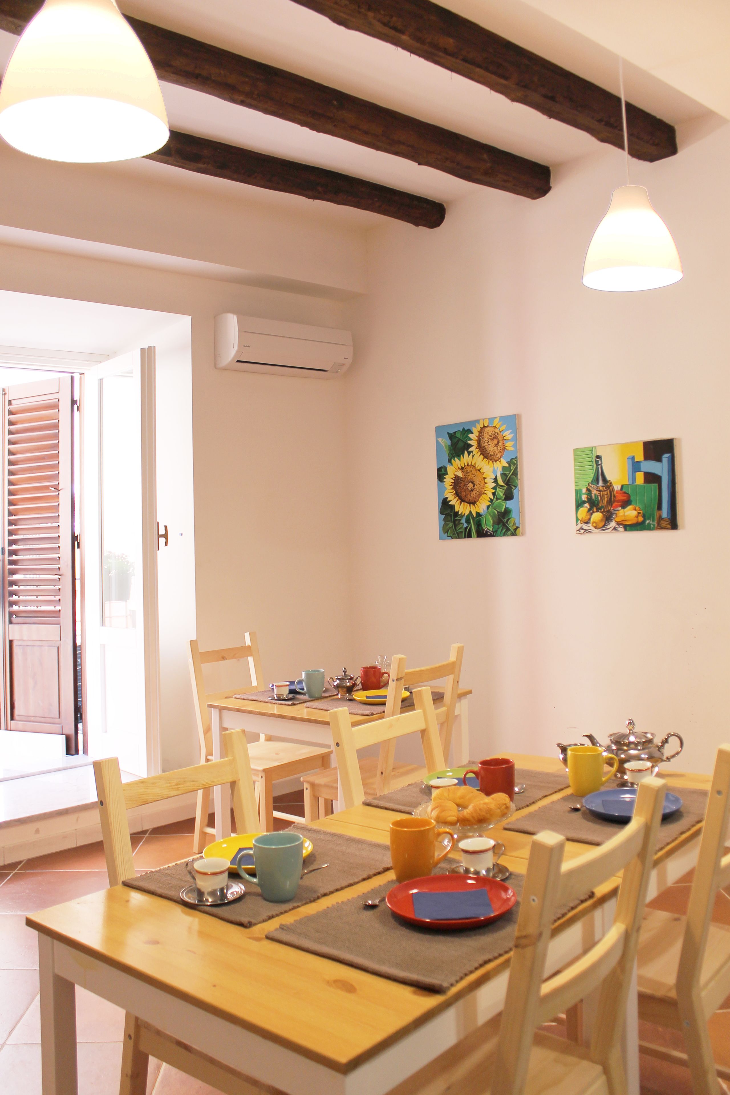 A dining area with wooden chairs and a table set for breakfast. There are two paintings on the wall, a window with blinds, and an air conditioning unit.