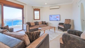 Un salon avec un canapé, des fauteuils, une table basse et un téléviseur. Il y a une fenêtre donnant sur la mer.