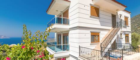 A multi-story white building with balconies, a staircase, and a view of the sea.