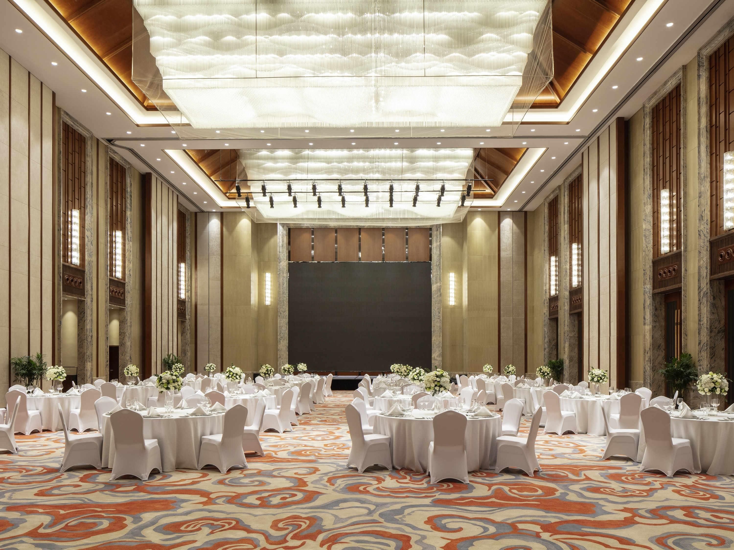 A banquet hall with round tables, white chairs, and a patterned carpet.
