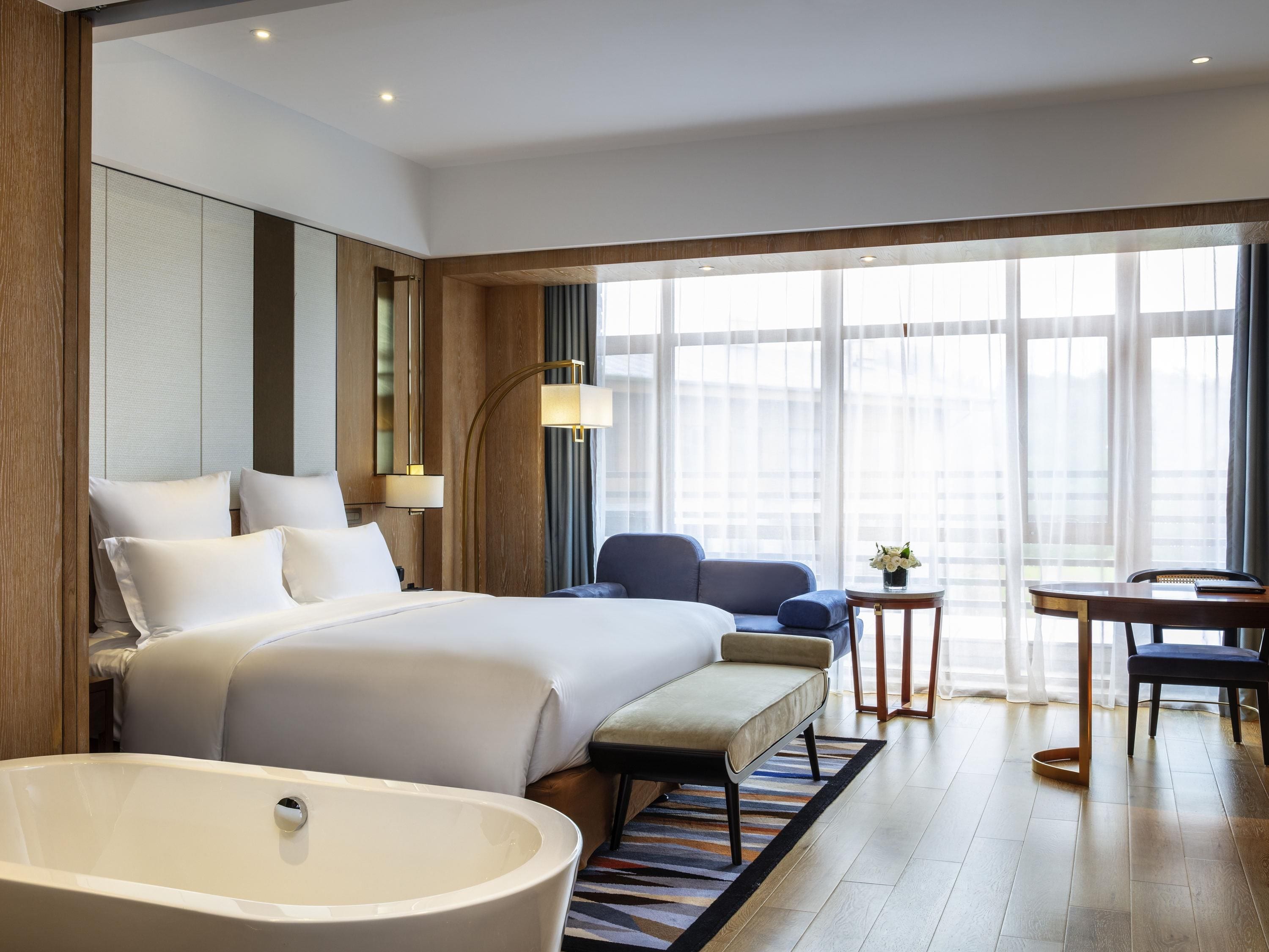 Una habitación de hotel moderna con una cama grande, una zona de estar, una mesita y una bañera.