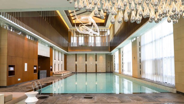 A spacious indoor swimming pool with a modern design, featuring a central seating area and a large ceiling light fixture. - Pullman Changbaishan Resort (Baishan)
