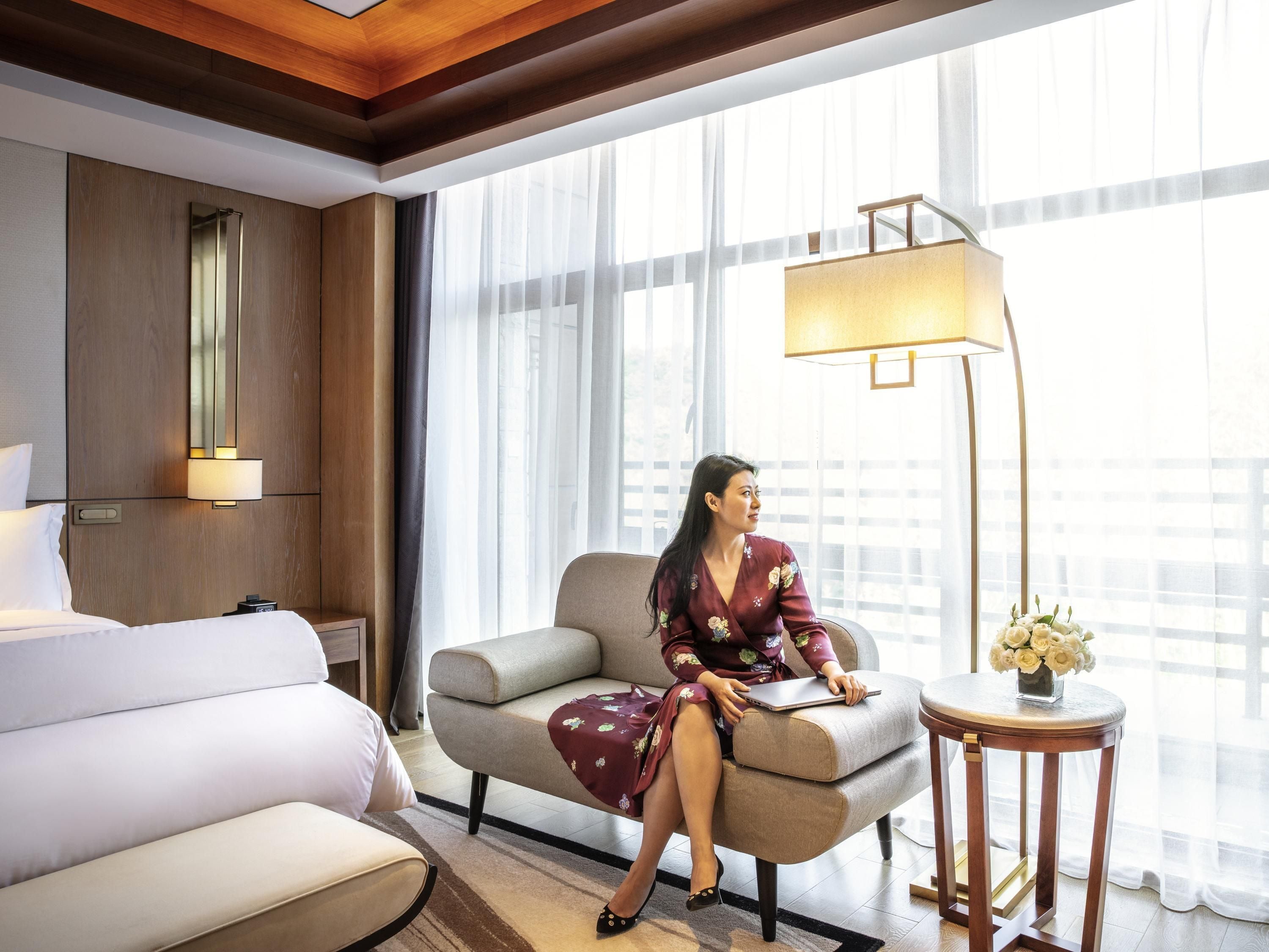 A hotel room with a bed, a sofa, a desk with a lamp, and a large window with sheer curtains.
