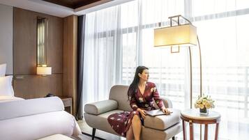 A hotel room with a bed, a sofa, a desk with a lamp, and a large window with sheer curtains.