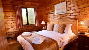 A wooden cabin bedroom with a large bed, two bedside tables with lamps, a window with curtains, and a framed picture on the wall. - Leny Estate (Callander)