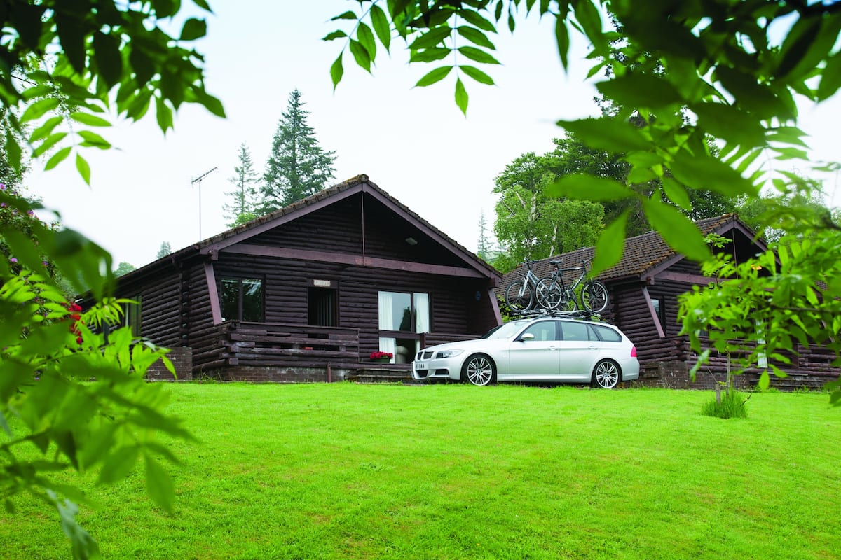 A rustic cabin with a car parked in front, surrounded by lush greenery.