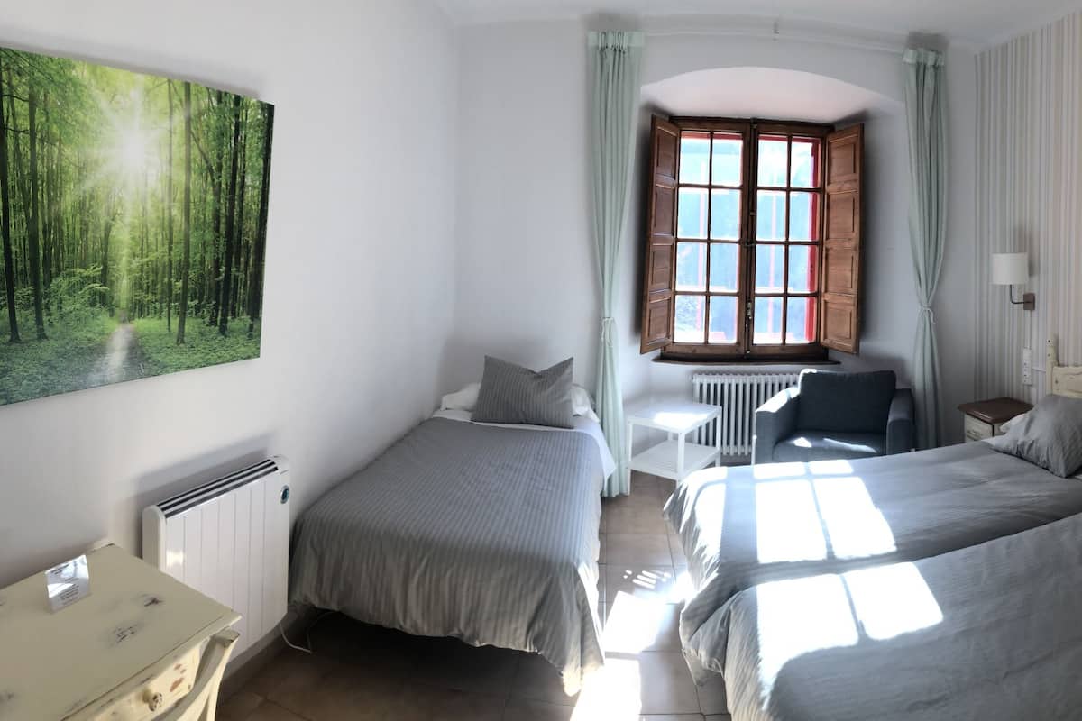 A bedroom with two beds, a radiator, a desk, a chair, a lamp, a window, and a painting of a forest.