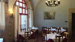 Un espace repas avec des tables dressées, des chaises en bois, un lustre et une grande fenêtre en arc de cercle dotée de montures rouges.
