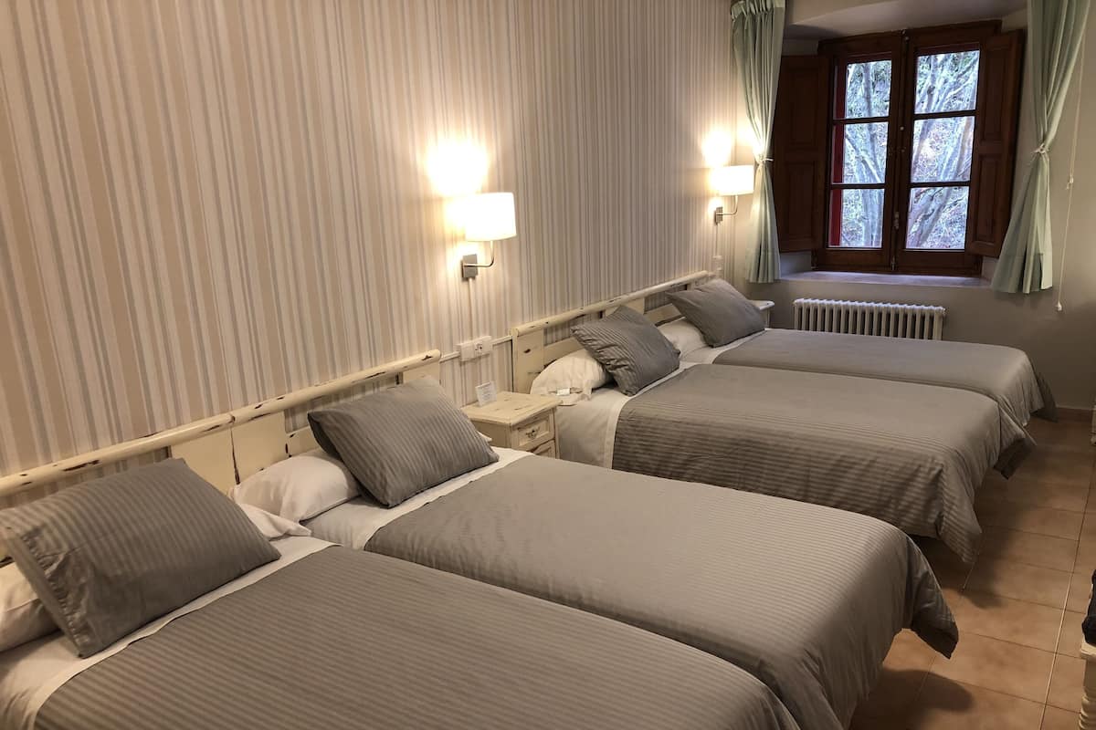 A hotel room with four single beds, a window with curtains, a radiator, and two wall-mounted lamps.