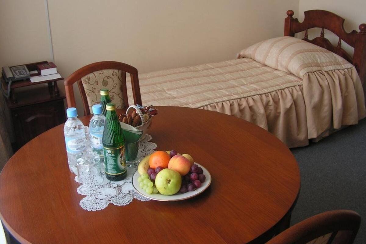 A room with a bed, a bedside table with a book and a phone, and a round table with a fruit bowl and water bottles.