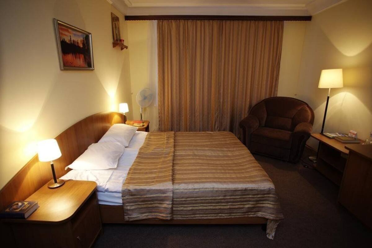 A hotel room with a bed, two bedside tables with lamps, a chair, a desk with a lamp, a fan, and a curtain.