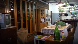 A compact kitchen with wooden cabinets, a small table with a floral tablecloth, and a hanging plant. - Baan Aunjai Homestay (Mae Lao)