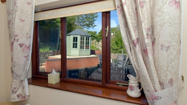 A window with a view of a green shed and a brick wall, curtains with a floral pattern, and a white vase on the windowsill. - Erris Villa (Builth Wells)