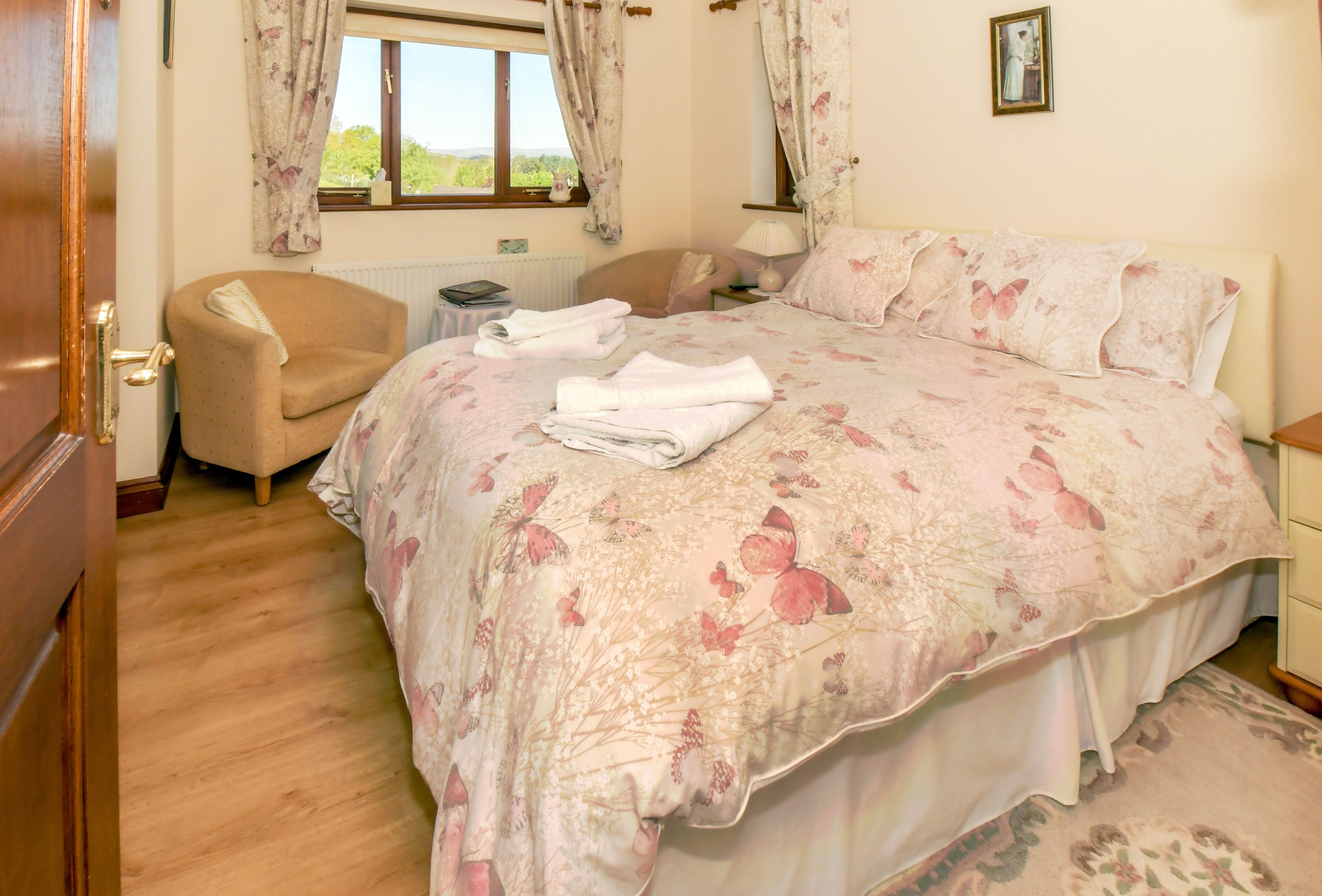 A bedroom with a floral bedspread, a chair, a bedside table with a lamp, and a window with curtains.