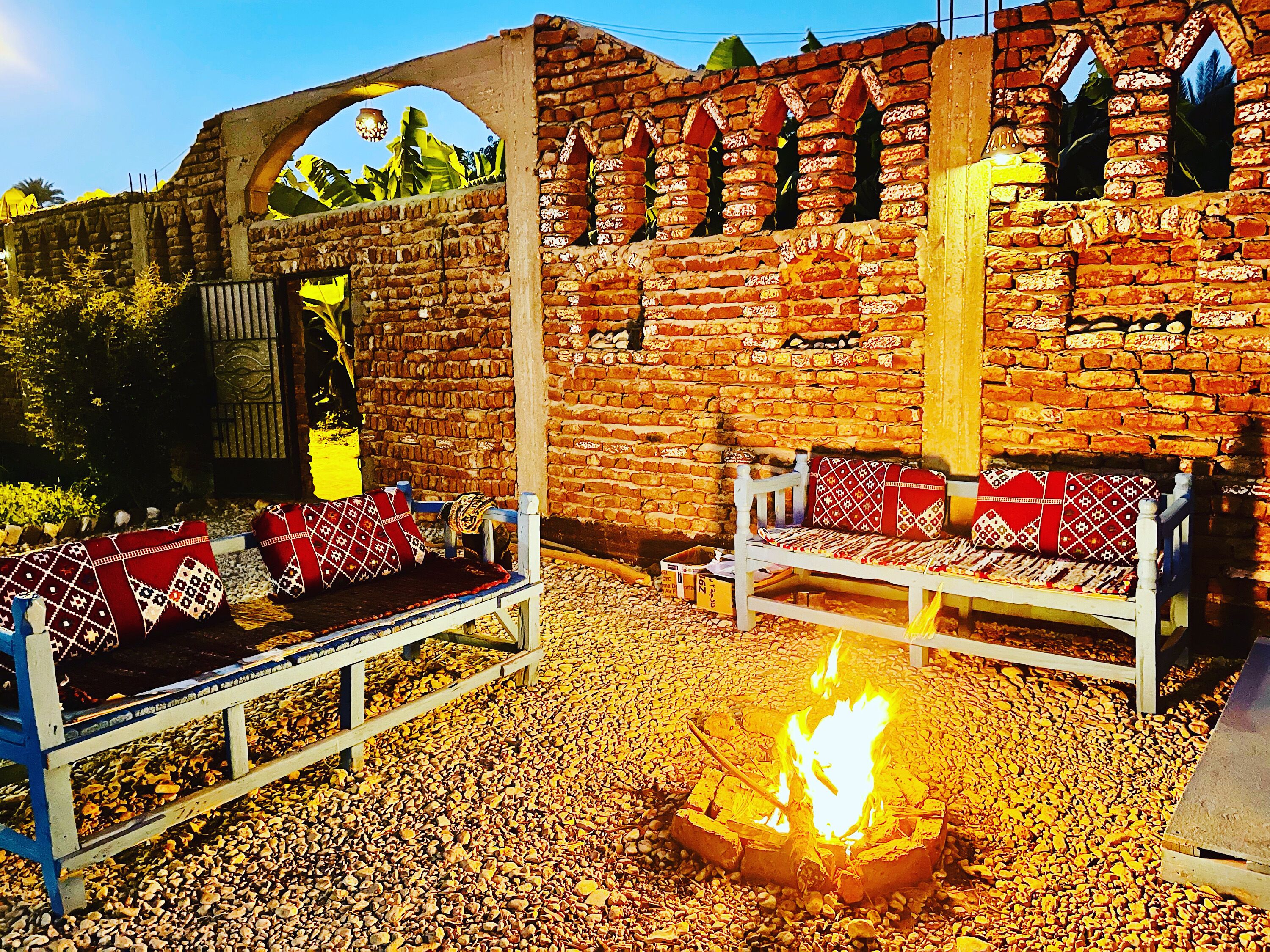 A rustic outdoor seating area with a fire pit, benches with patterned cushions, and a brick wall backdrop.