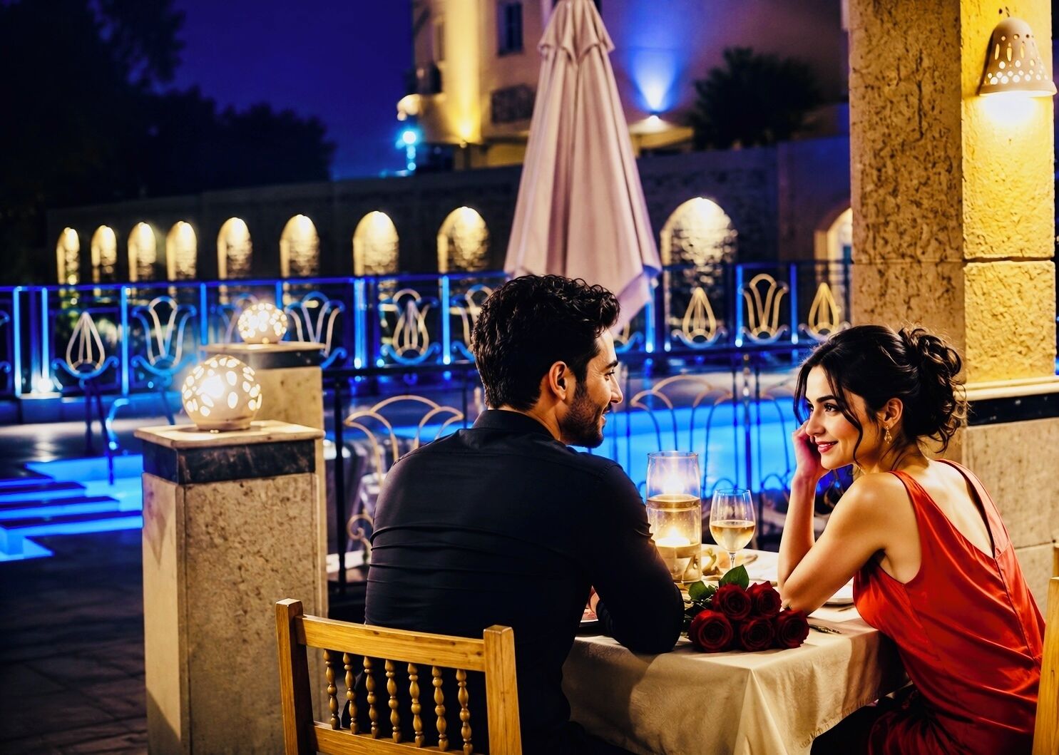 A couple dining at an outdoor table with a view of a pool and decorative lights.