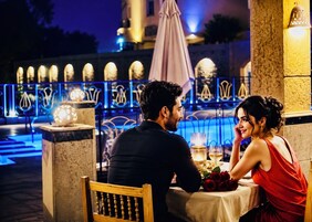 A couple dining at an outdoor table with a view of a pool and decorative lights.