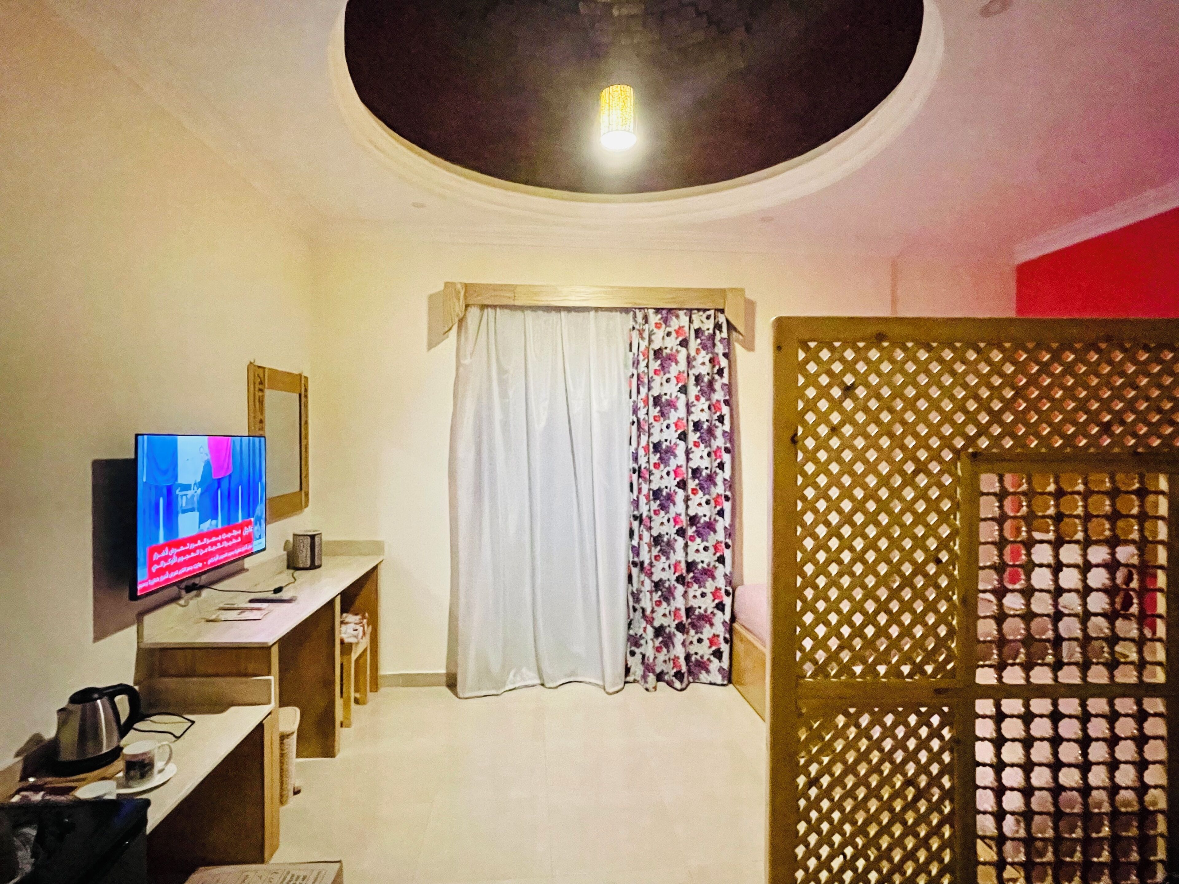 A hotel room with a television, a desk with a kettle and cups, a bed with a white and floral curtain, and a decorative lattice partition.