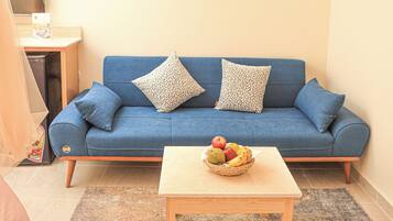 A living room with a blue sofa, two patterned cushions, a wooden coffee table with a fruit bowl, and a painting on the wall.