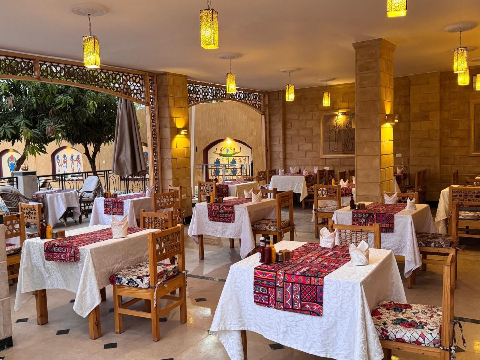Un restaurante con mesas cubiertas de manteles blancos y caminos de mesa con estampado rojo, sillas de madera y lámparas colgantes con diseño de farol.