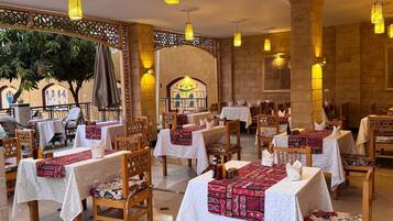 Un restaurante con mesas cubiertas de manteles blancos y caminos de mesa con estampado rojo, sillas de madera y lámparas colgantes con diseño de farol.