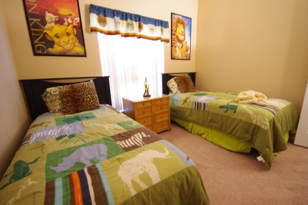 Quarto com duas camas, cada uma com colcha de tema animal diferente. Há duas pinturas na parede, uma de leão e outra do Rei Leão. Um penteadeira de madeira fica entre as camas, e uma janela com cortinas é visível.