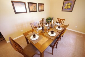 A dining room with a wooden table set for four, surrounded by chairs. There are framed pictures on the wall, a potted plant, and a vase on the table.