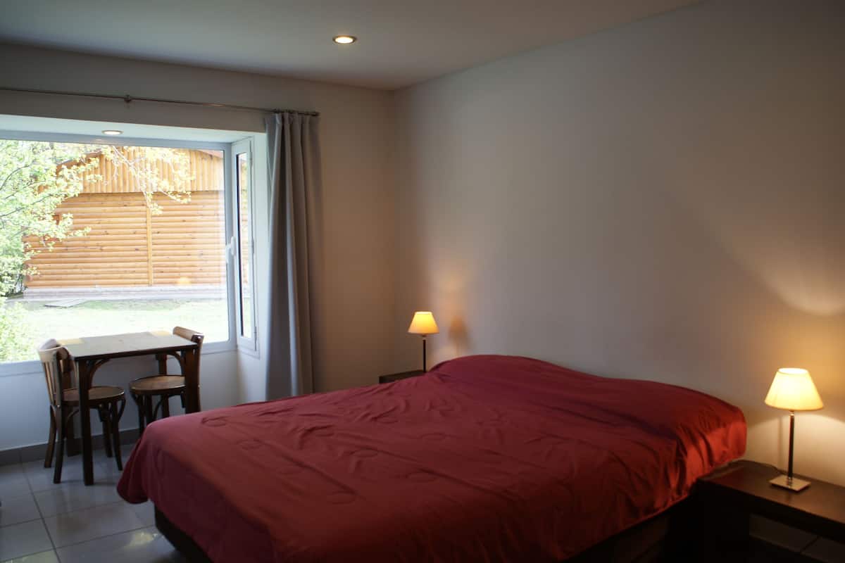 A bedroom with a bed, two bedside lamps, a window with curtains, a table with chairs, and a view of a wooden fence outside.