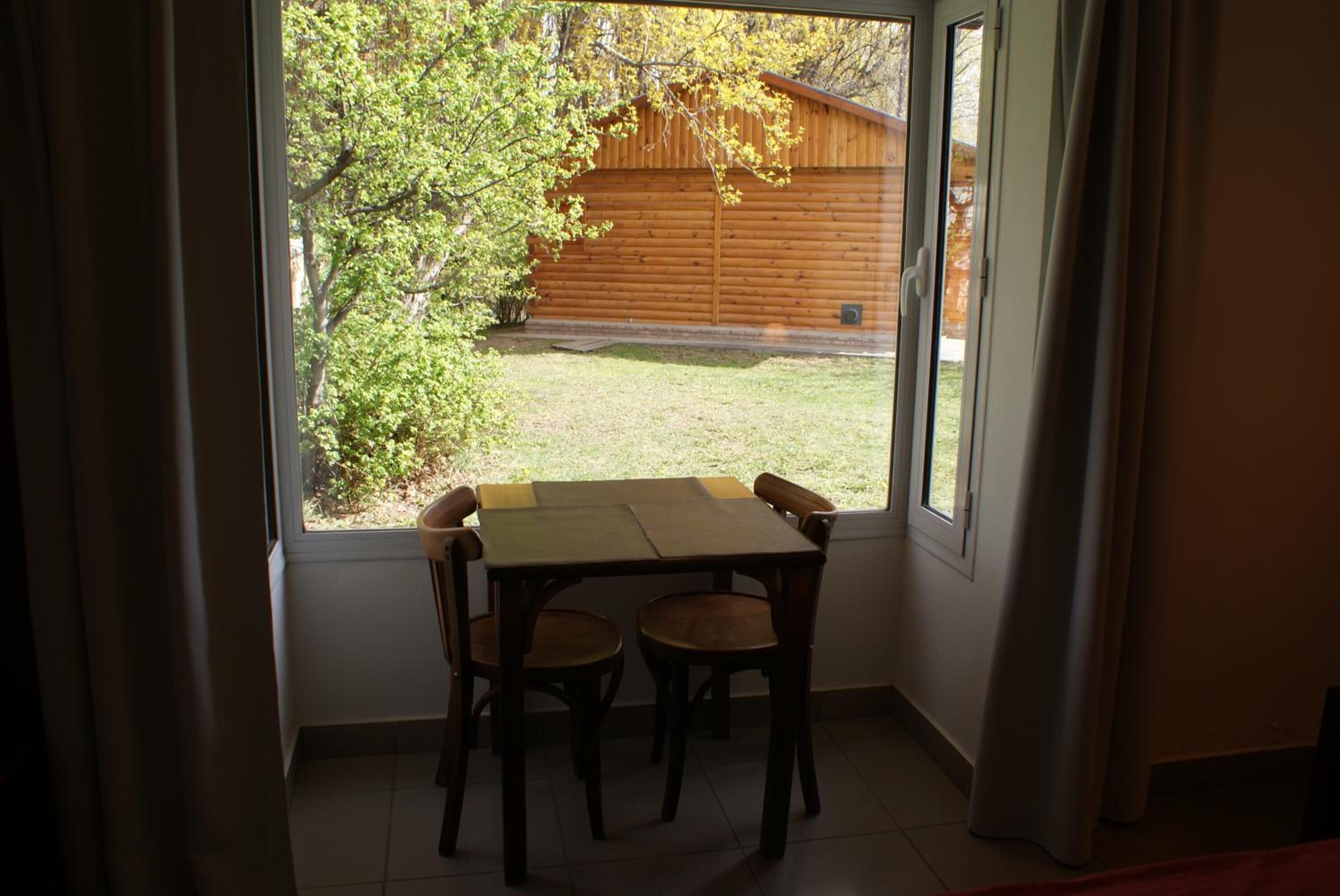 A room with a table and chairs, a window with curtains, and a view of a wooden fence and trees.