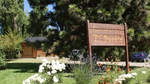Un letrero de madera con la inscripción 'Cabanas El Puente', rodeado por un jardín con diversas flores y un edificio al fondo.