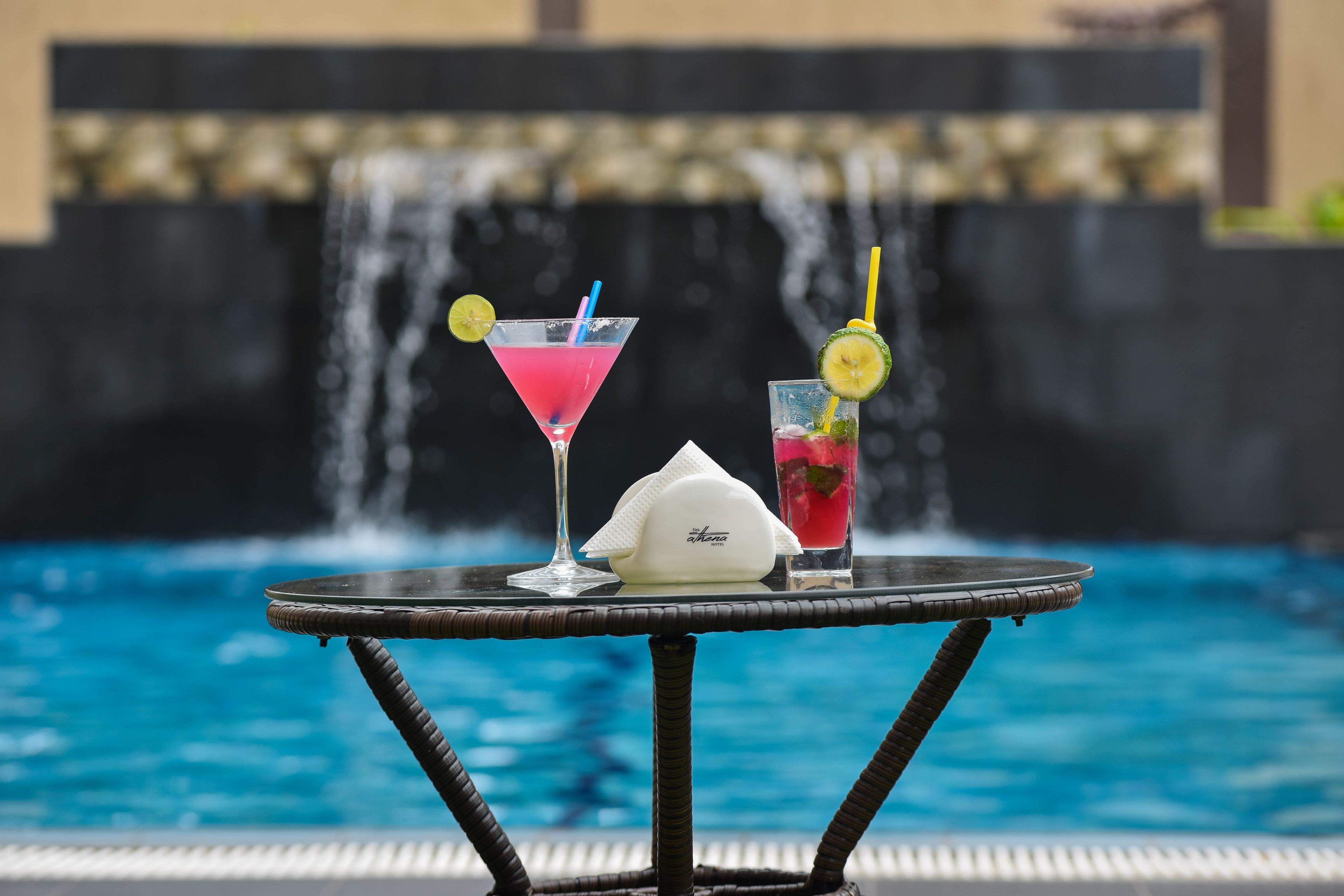 A table with two drinks and a napkin by a pool with a waterfall backdrop.