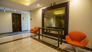 Un vestíbulo de hotel con piso de mármol, un mostrador de recepción, dos sillones naranja y un espejo grande.