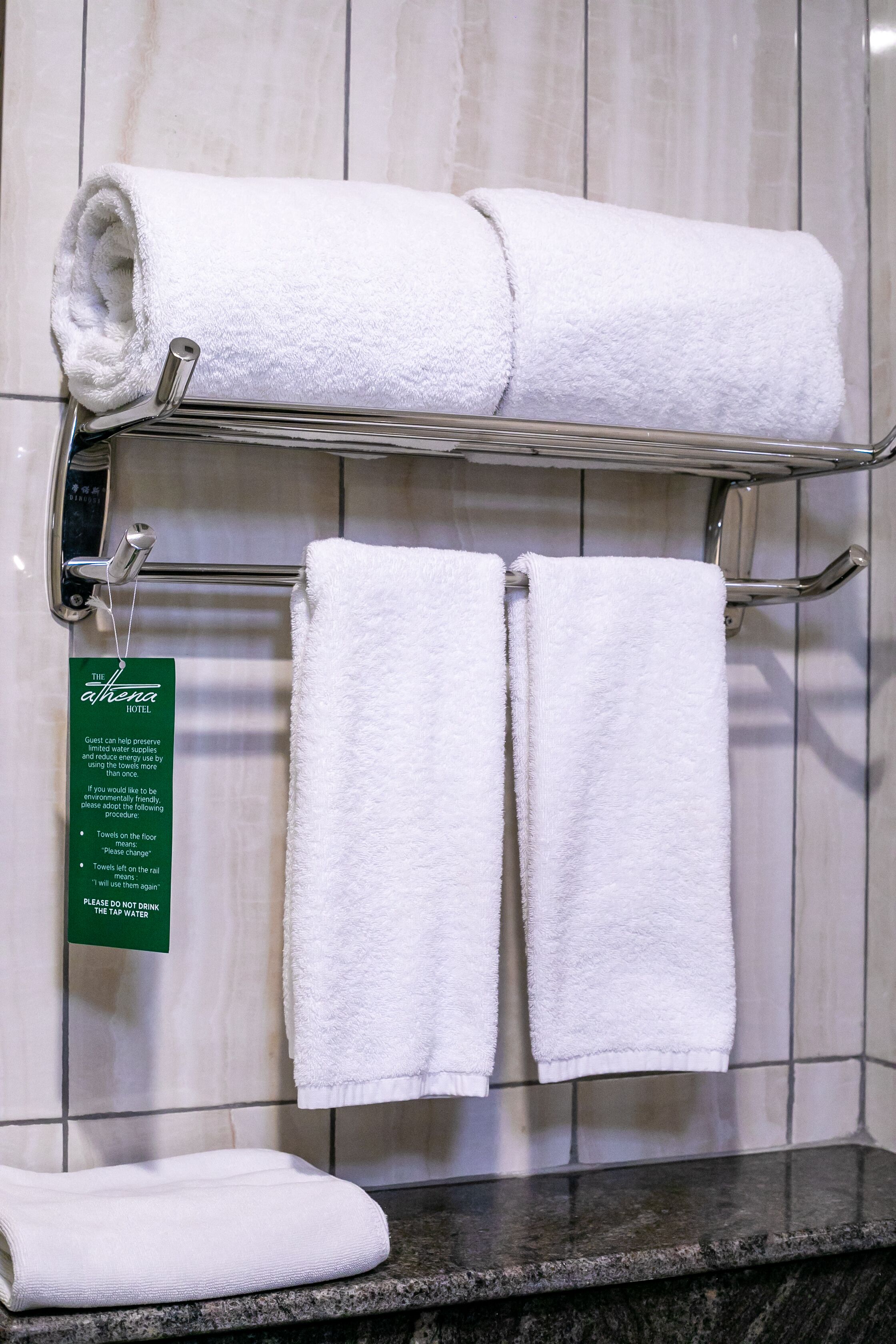 Ein Hotelbad mit einem Handtuchhalter, der drei weiße Handtücher trägt, einem grünen Schild mit Hotelinformationen und einer Marmorarbeitsplatte.