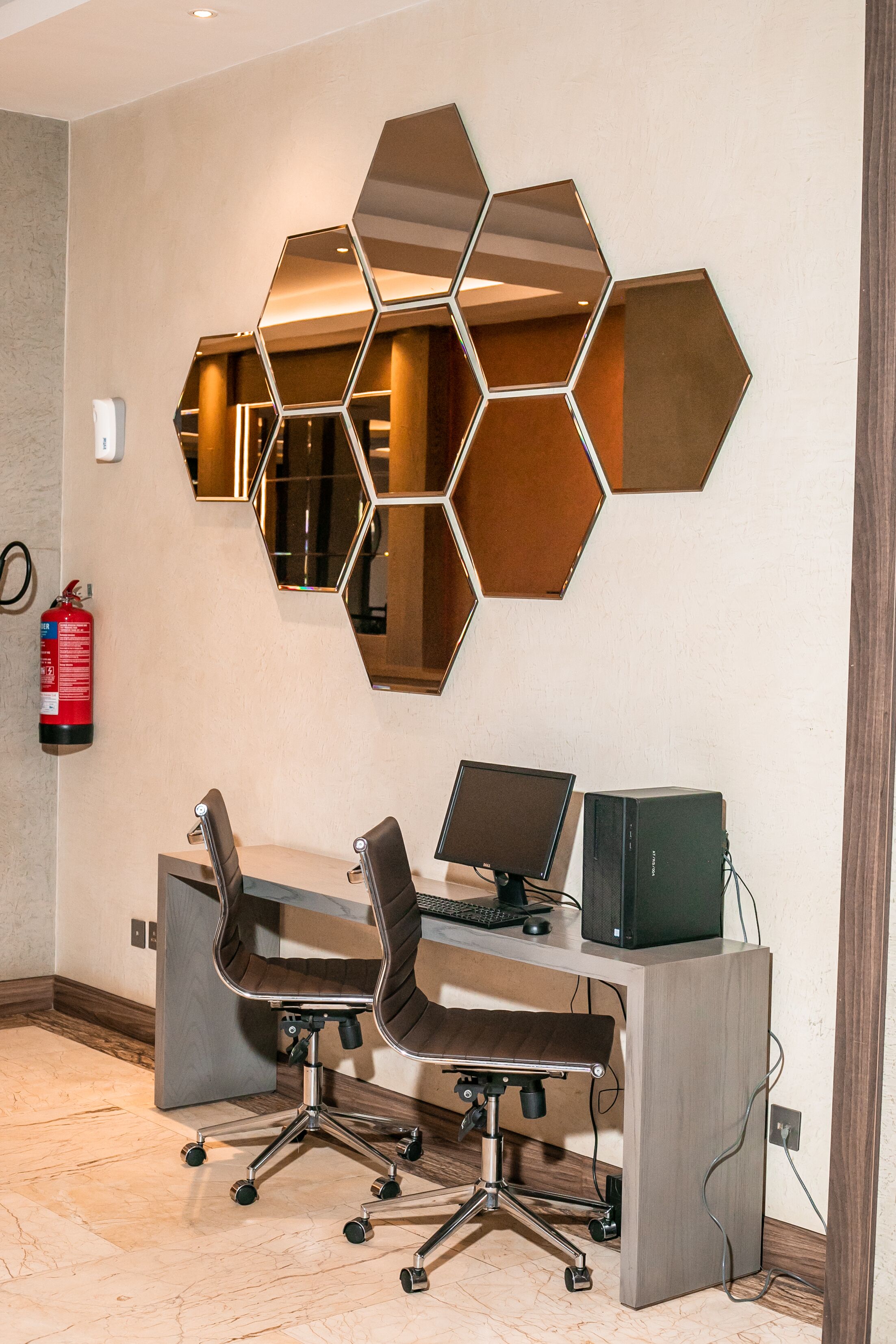 Um espaço de escritório moderno com um painel de espelhos hexagonais, duas cadeiras de escritório, uma escrivaninha com um computador e um extintor de incêndio na parede.