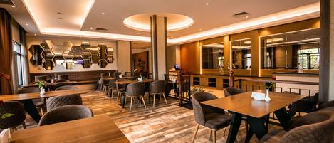 A modern restaurant with wooden tables and chairs, a bar area, and a seating section with a high ceiling and recessed lighting.
