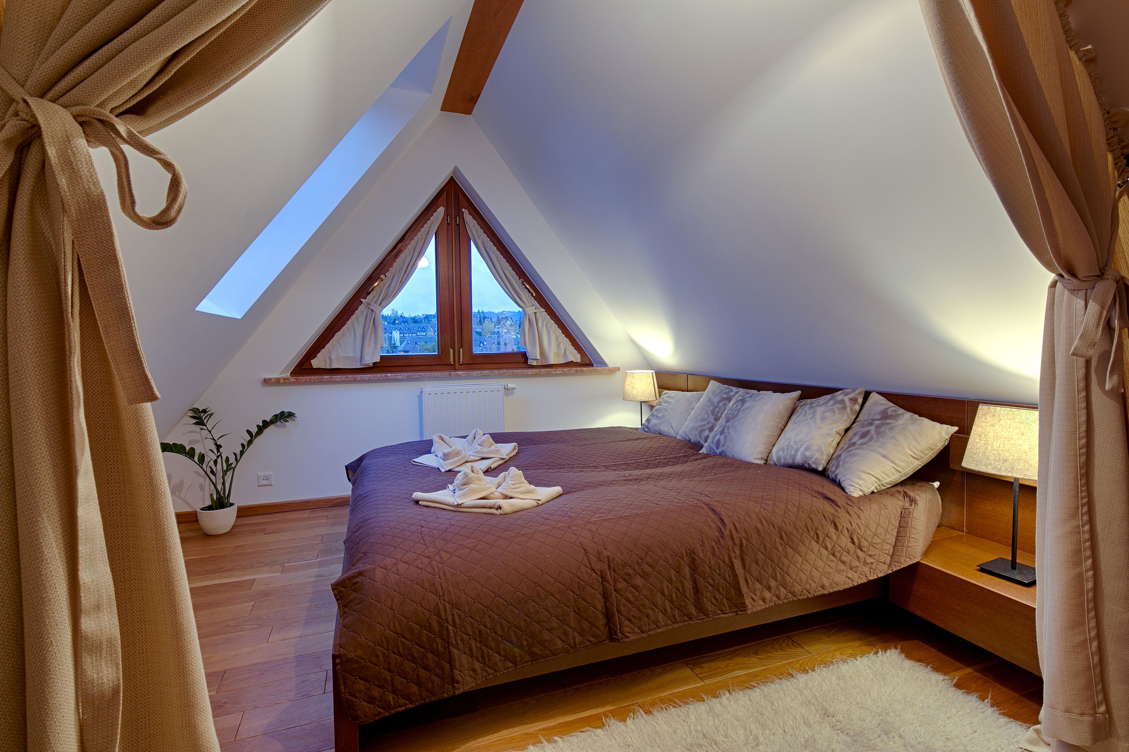A bedroom with a sloped ceiling, a large window, a bed with a brown quilt, a bedside table with a lamp, and a potted plant.