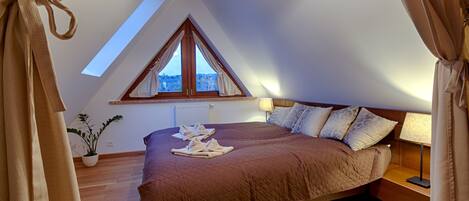 A bedroom with a sloped ceiling, a large window, a bed with a brown quilt, a bedside table with a lamp, and a potted plant.