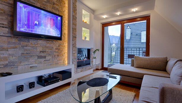 A modern living room with a flat-screen TV mounted on a stone wall, a glass coffee table, a beige sofa, and a fireplace. - Apartament Ogrody Górskie widok na Tatry (Zakopane)