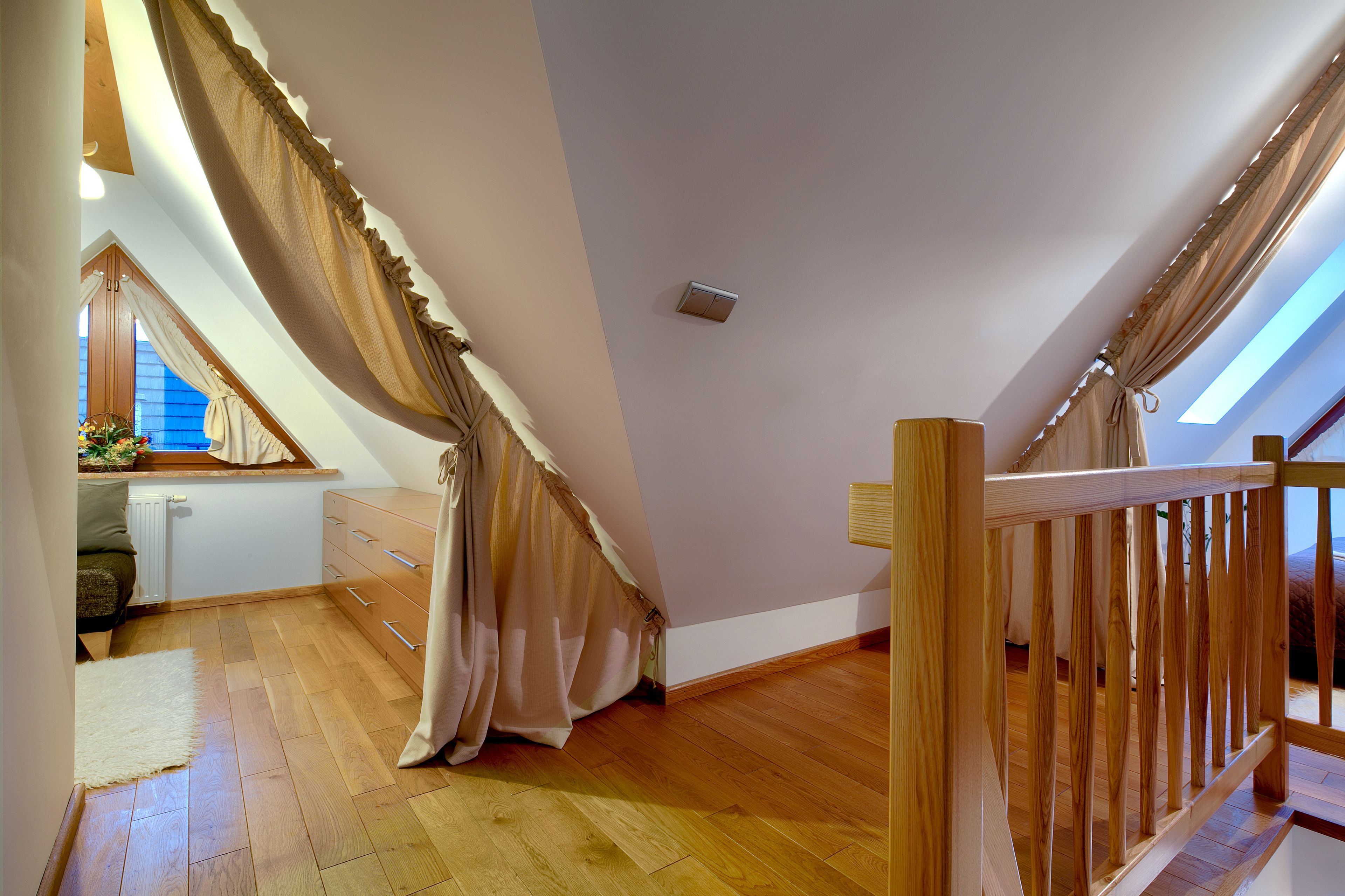 Una habitación con techo inclinado, suelo de madera, ventana con cortinas, radiador y barandilla de madera.