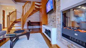 A modern living room with a wooden staircase, a flat-screen TV mounted on a stone wall, and a fireplace. - Apartament Ogrody Górskie widok na Tatry (Zakopane)